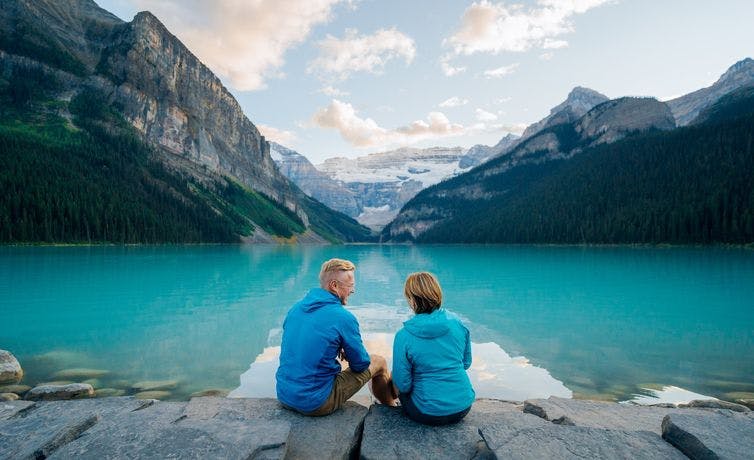 A mature couple sits on the rocks at the edge of a vast turquoise blue lake with a glacier and large mountains in view at the end of the lake