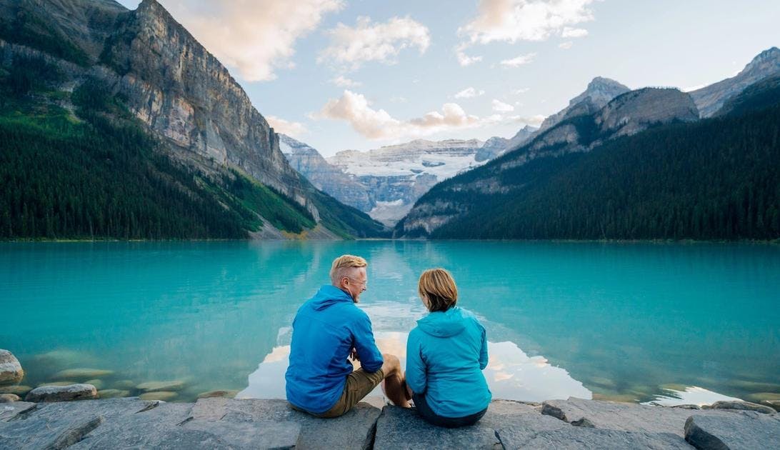 A mature couple sits on the rocks at the edge of a vast turquoise blue lake with a glacier and large mountains in view at the end of the lake
