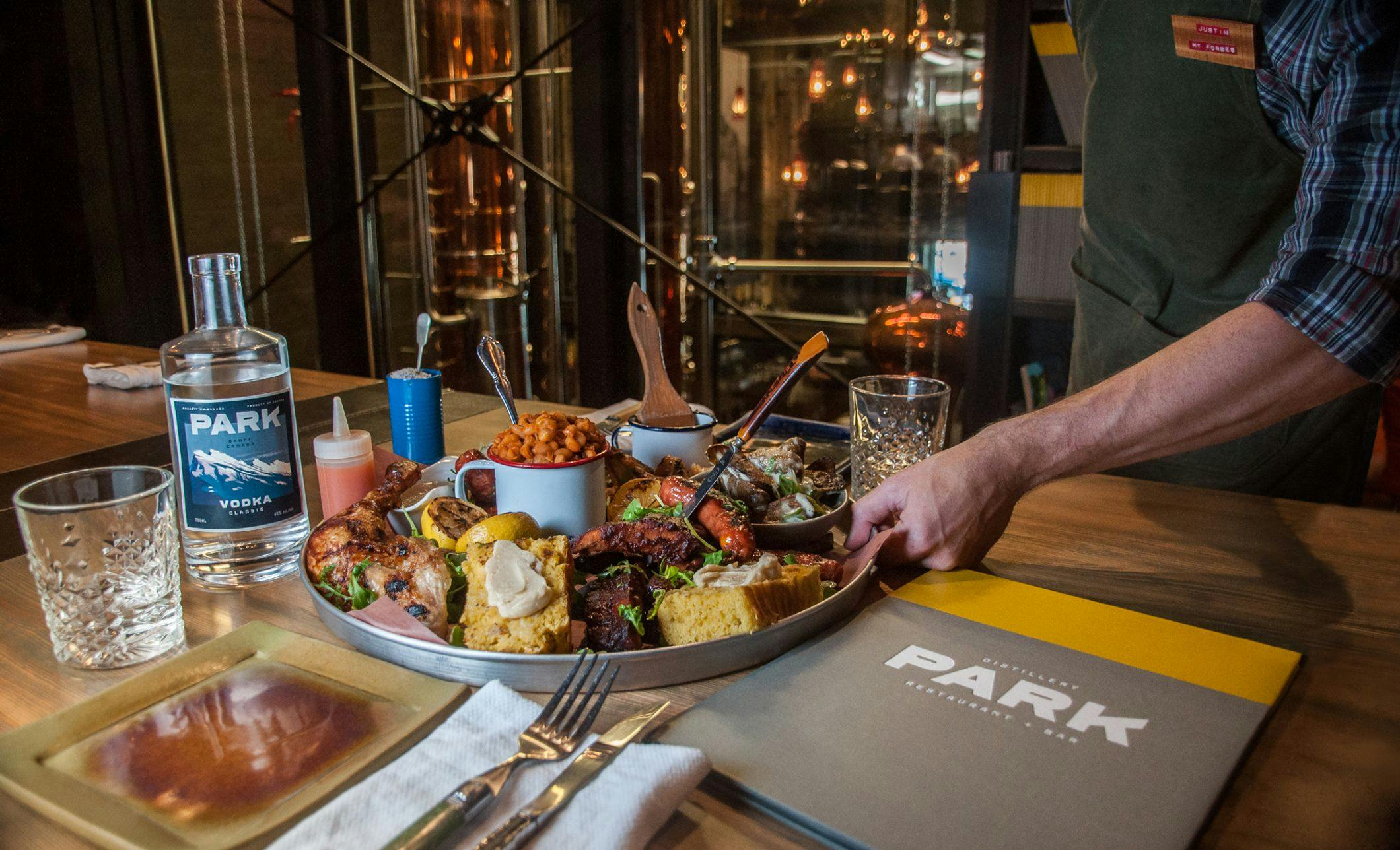 A server lays down a large sharing plat of food with a bottle of vodka and the distillery in the background