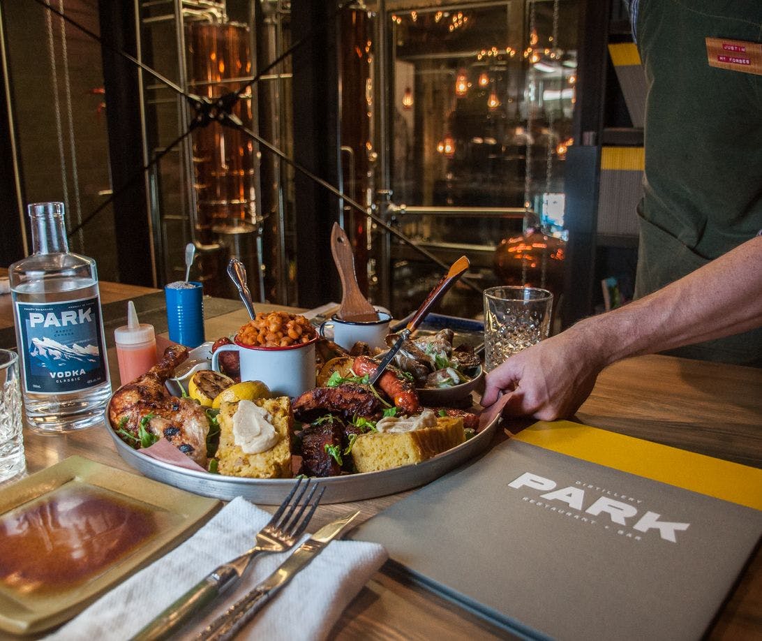 A server lays down a large sharing plat of food with a bottle of vodka and the distillery in the background