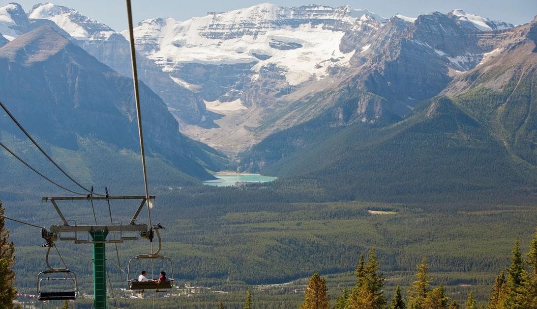 A couple travels down a mountain in an open chairlift while taking in the view of Lake Louise and Victoria Glacier in the distance